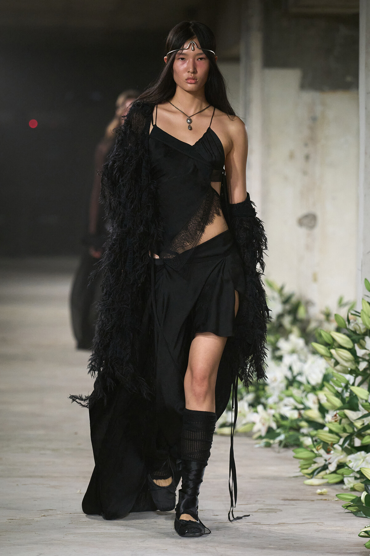 Model walks a runway in a black, asymmetrical dress with lace panels and a long feathered coat, paired with knee-high boots.