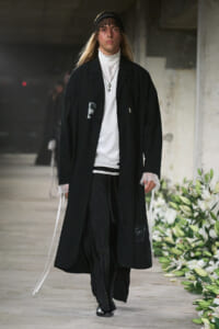 Male model walks a runway in an oversized black coat over a white high-neck top, with lace cuffs and layered necklaces, in an industrial setting with floral decor.