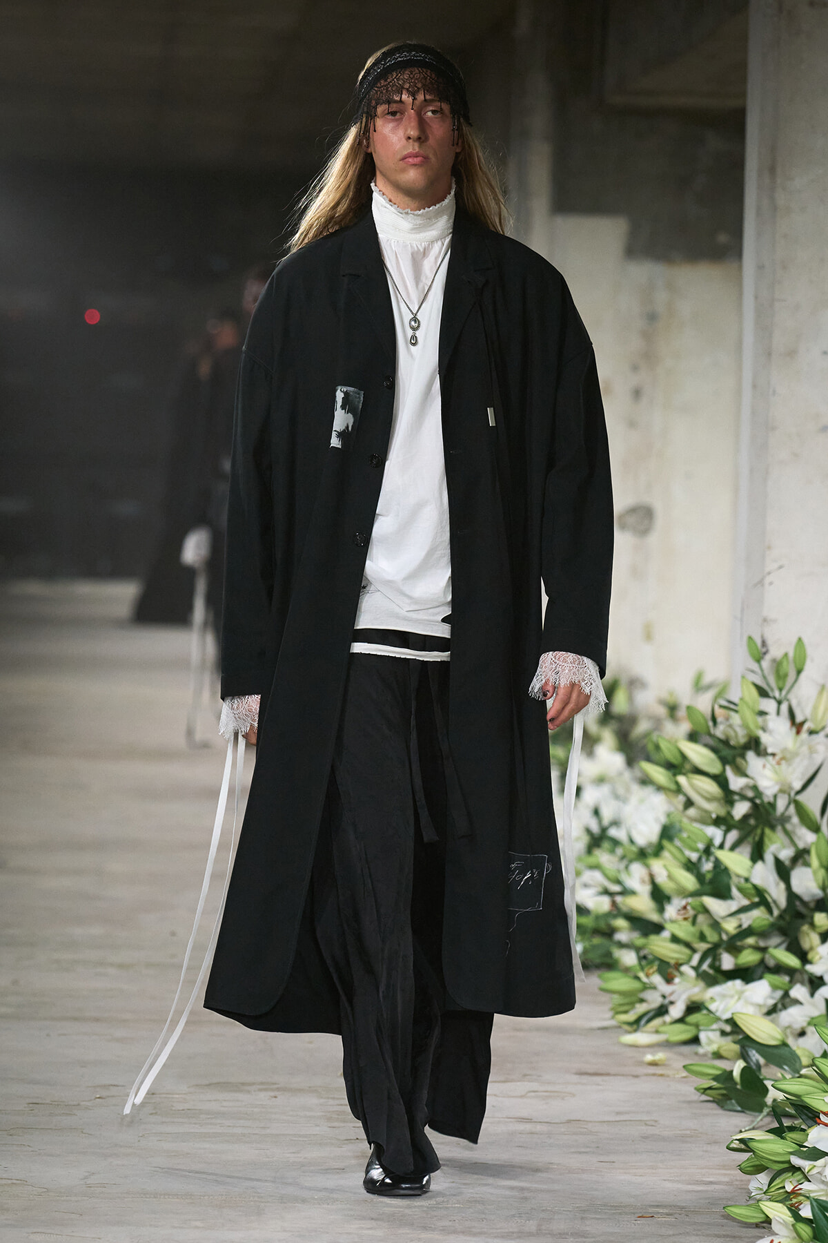 Male model walks a runway in an oversized black coat over a white high-neck top, with lace cuffs and layered necklaces, in an industrial setting with floral decor.