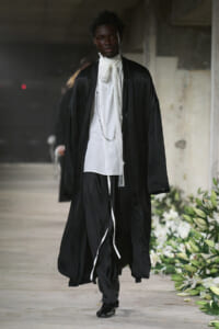 Male model on a runway wearing a long black robe over a white shirt with a cravat and hanging chains, in an industrial venue.