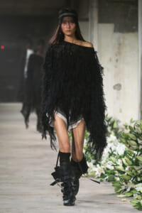 Model walking a runway in an oversized black feathered top, off-shoulder, paired with chunky black boots and a lace headband.
