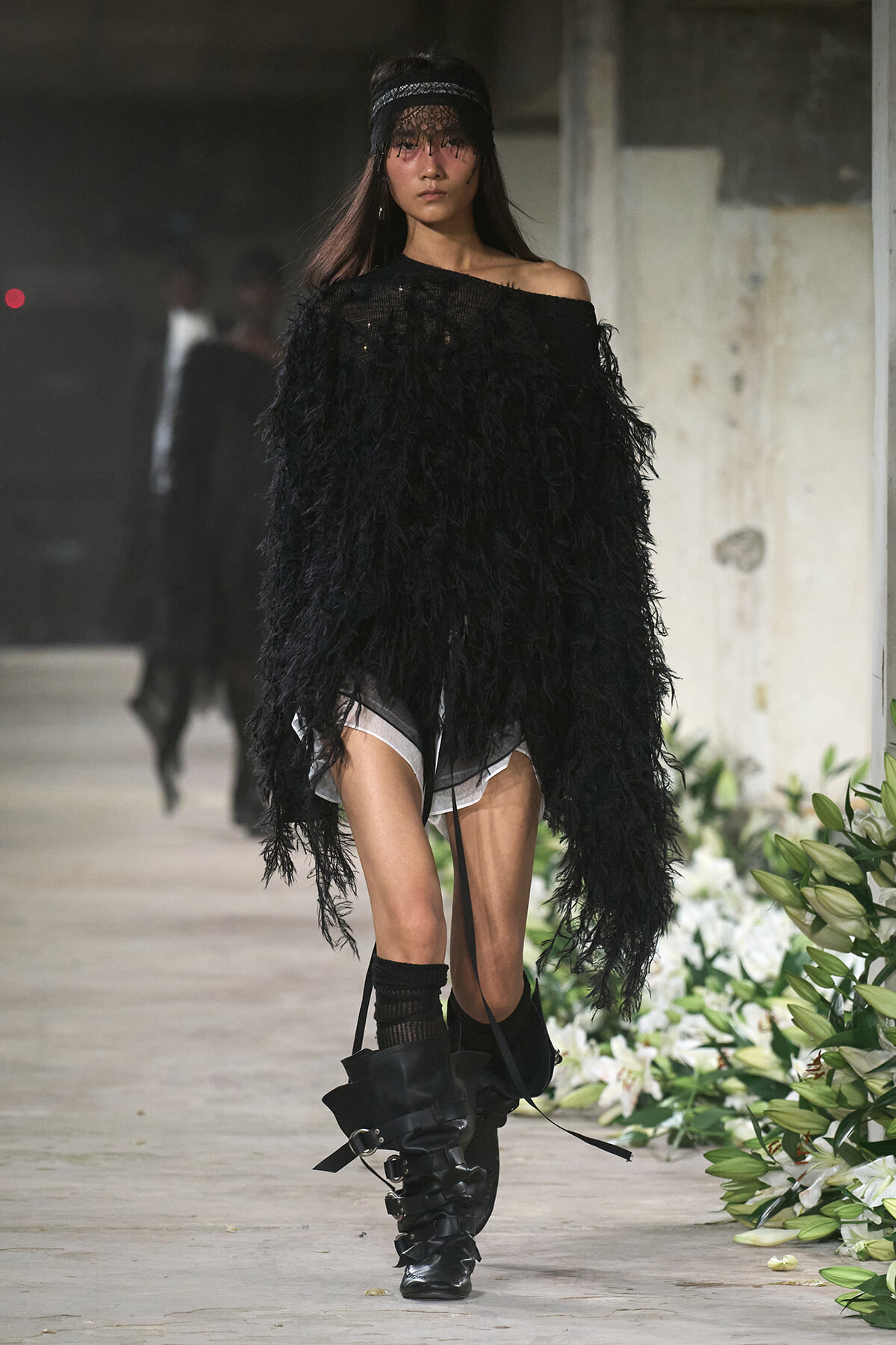 Model walking a runway in an oversized black feathered top, off-shoulder, paired with chunky black boots and a lace headband.