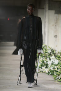 Model walking a runway in a black sheer Victorian-inspired gown with lace cuffs, layered necklaces, and white sneakers.