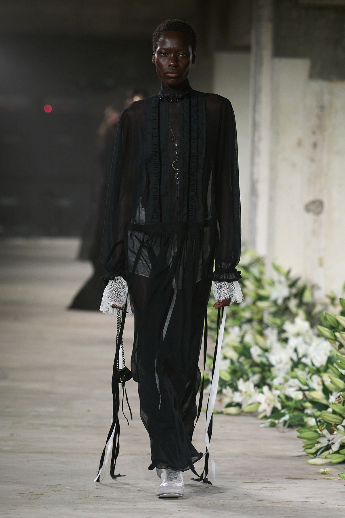 Model walking a runway in a black sheer Victorian-inspired gown with lace cuffs, layered necklaces, and white sneakers.