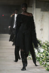 Model walking runway in all-black ensemble with feathered off-the-shoulder top and flowing sheer skirt, paired with tall layered boots.