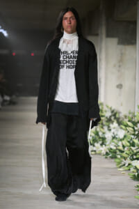 Runway model wearing an all-black, layered look with a white lace high-neck collar and a white graphic tee underneath.