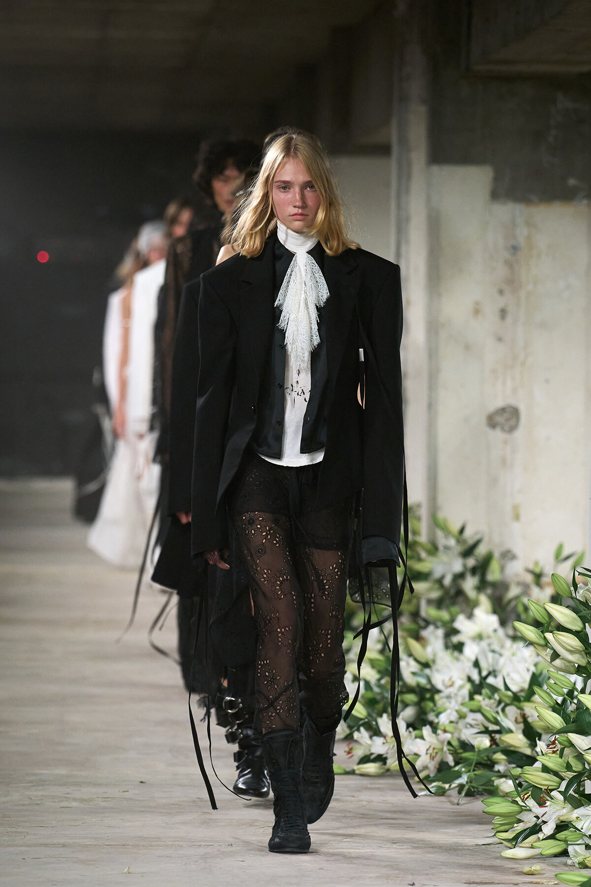 Model walking a fashion runway in a black blazer, white lace necktie, sheer patterned tights, and black boots.