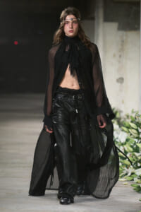 Model on a runway wearing sheer black cape over a cropped top and black leather pants, with a silver headband