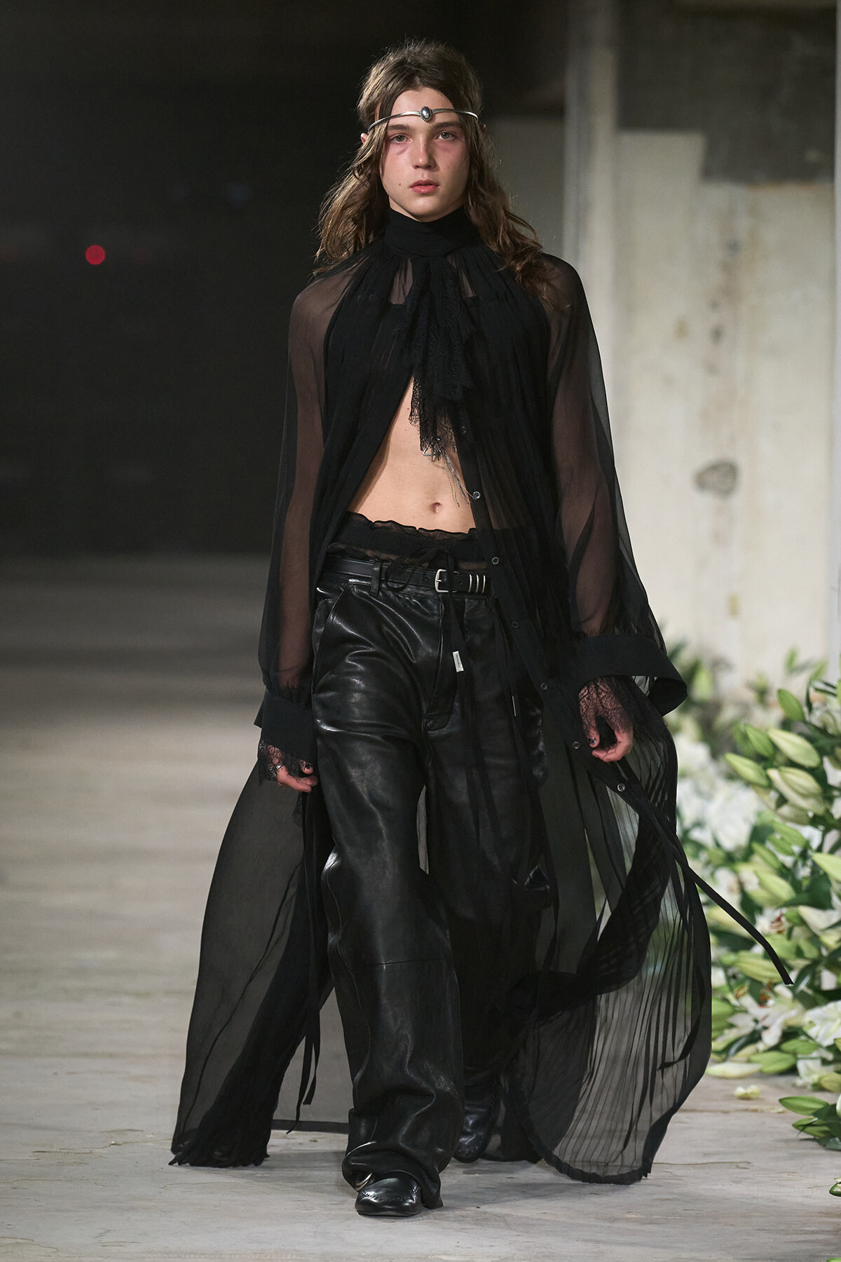 Model on a runway wearing sheer black cape over a cropped top and black leather pants, with a silver headband