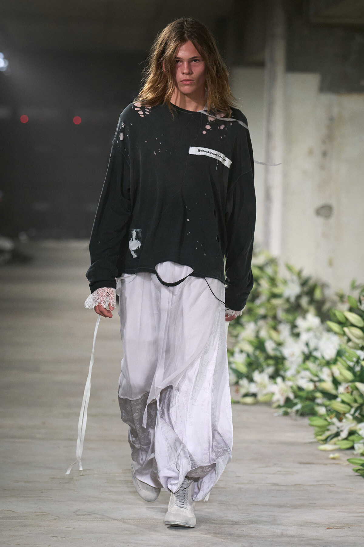 Model walking a runway in an oversized black distressed shirt and flowing white pants in an industrial venue with floral arrangements nearby.