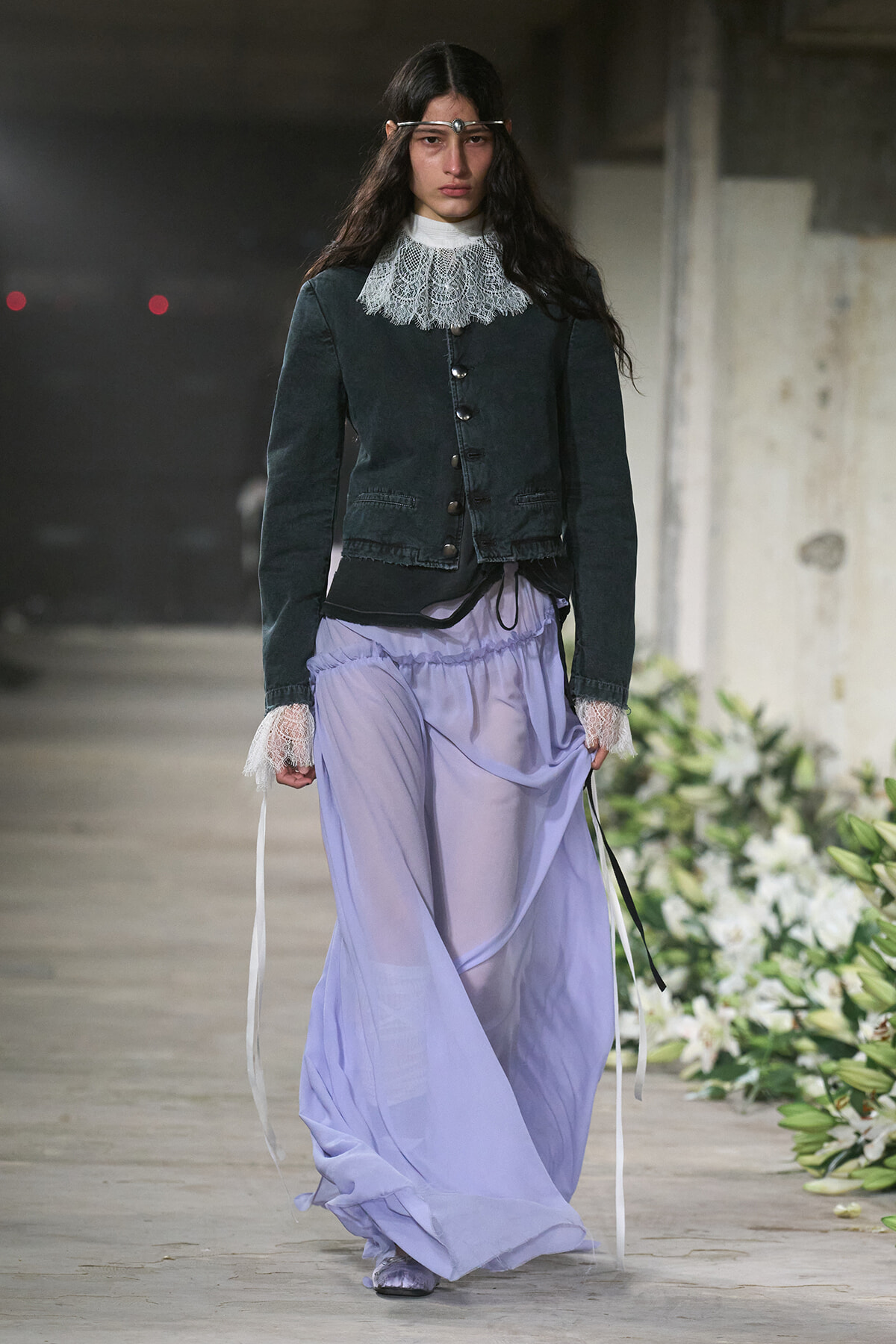 Model walking a runway in a dark green buttoned jacket with a lace collar, paired with a long lilac sheer skirt and lace-trim cuffs.