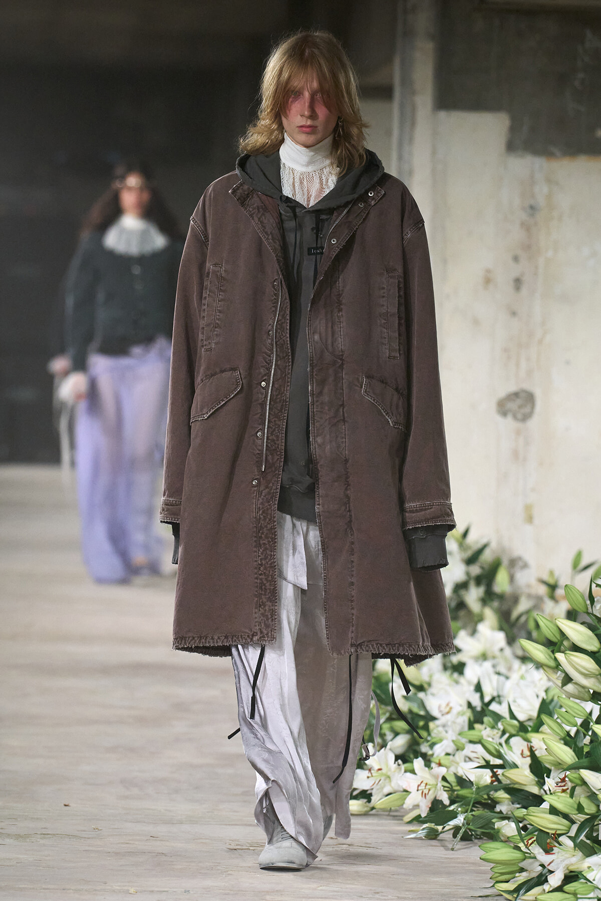 Model walking a runway in a oversized brown coat over layered black hoodie and light gray pants, industrial venue with white lilies along the catwalk