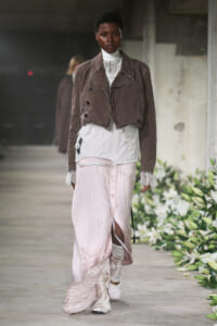 Model walking a runway in a brown cropped jacket over a white lace turtleneck, paired with a pale pink pleated skirt and layered underskirt, white socks, and sneakers amidst floral decor.