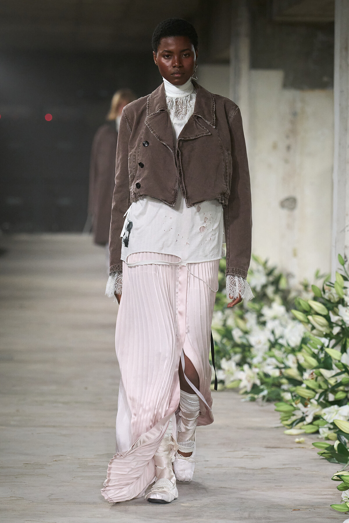 Model walking a runway in a brown cropped jacket over a white lace turtleneck, paired with a pale pink pleated skirt and layered underskirt, white socks, and sneakers amidst floral decor.
