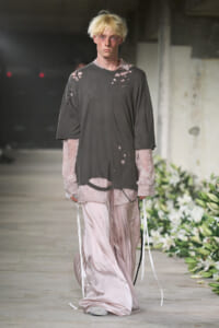 Male model wearing an oversized charcoal distressed shirt over pale pink lace sleeves and flowing satin pants on a runway