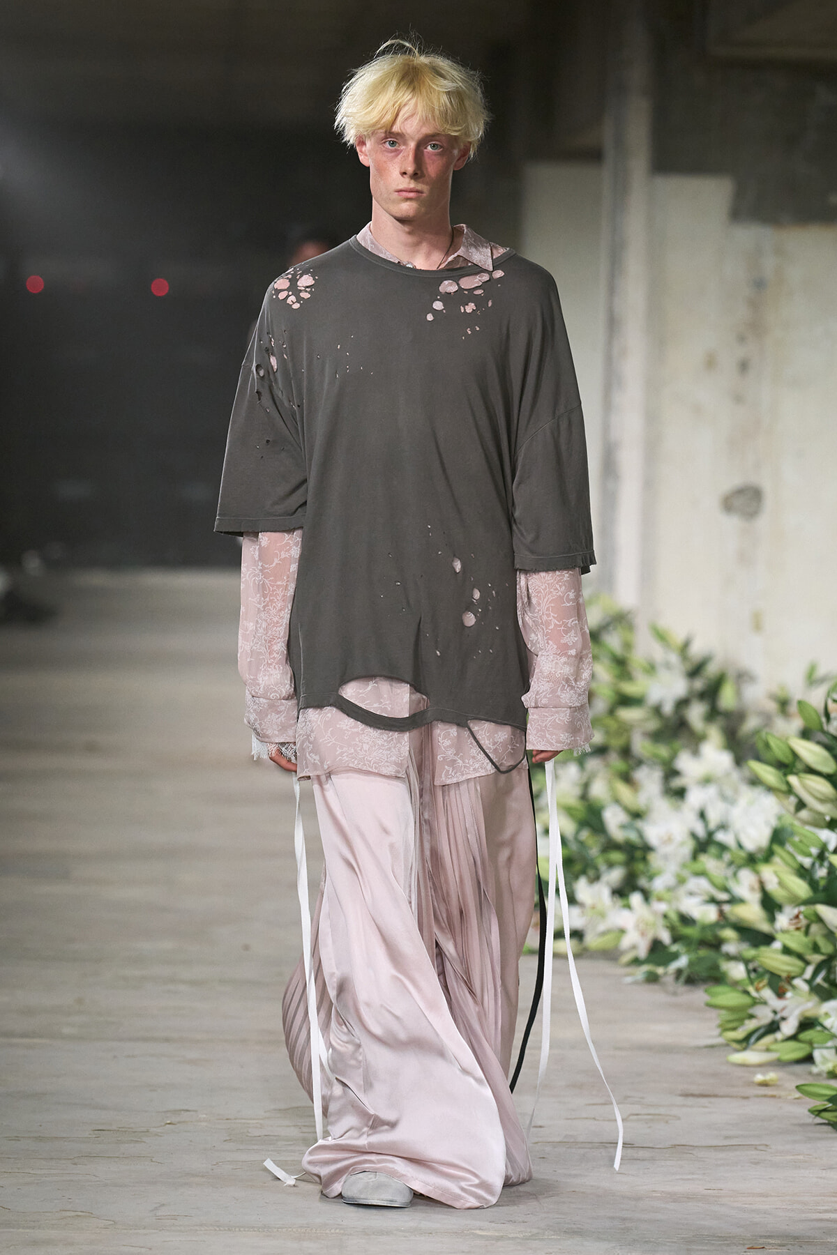 Male model wearing an oversized charcoal distressed shirt over pale pink lace sleeves and flowing satin pants on a runway