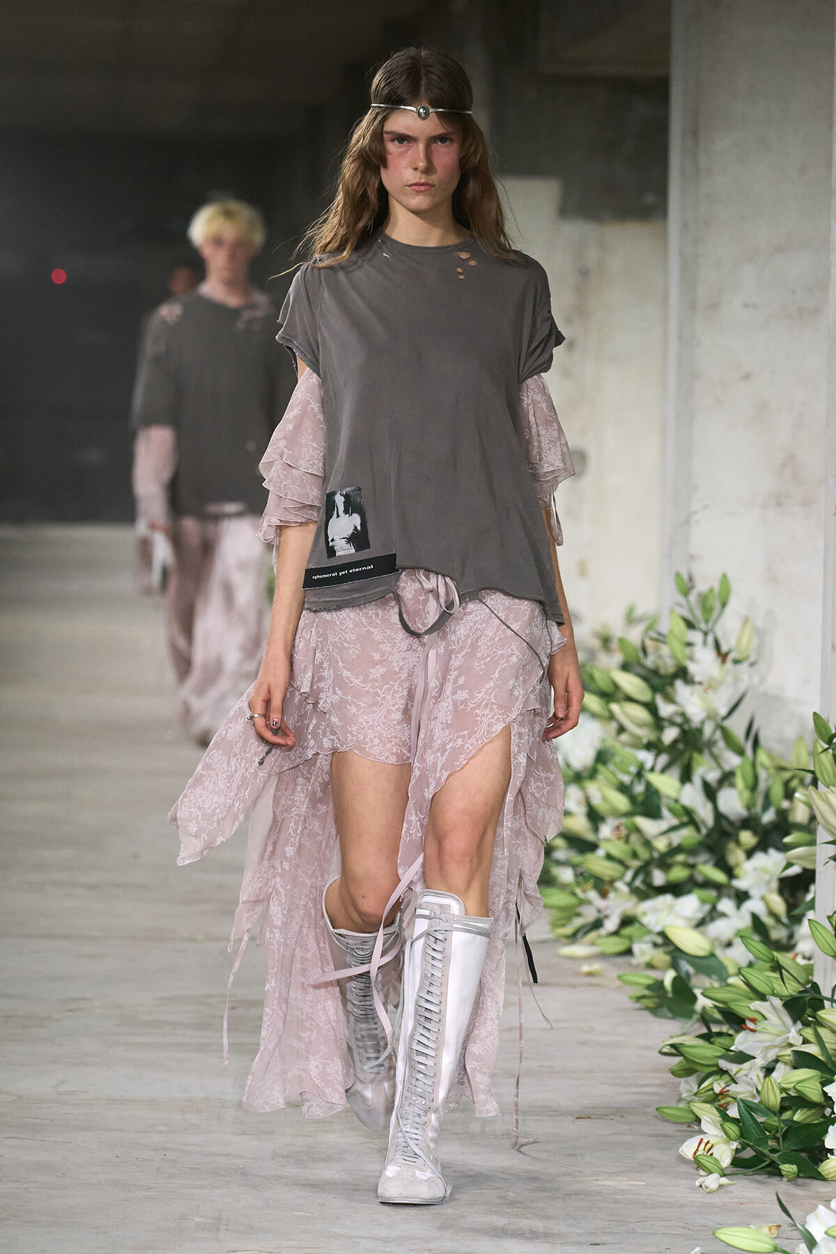 Model walking a fashion runway in a loose gray T-shirt over a pale pink lace dress with sheer layered sleeves, white knee-high lace-up boots, and a silver headband.
