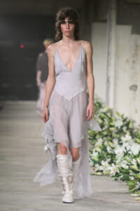Model walks a runway in a pale, sheer V-neck slip dress with a layered, asymmetrical skirt and white wrapped boots.