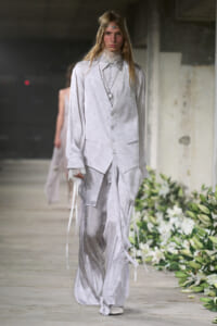 Model walking a runway in a pale silver-white, floral-patterned suit with a long vest-like jacket and flowing pants, accessorized with a headband and layered necklaces.