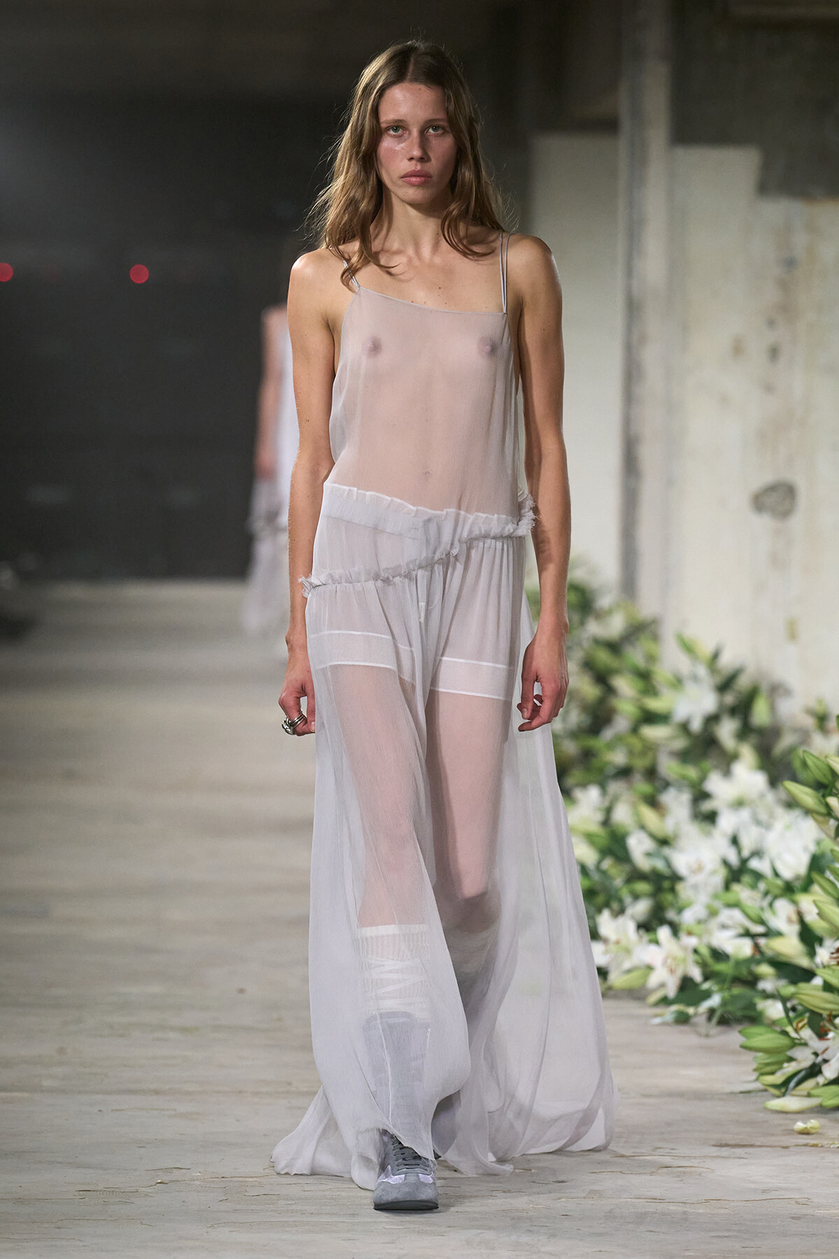 Model walks down a runway in a sheer, pale gray dress with thin straps and a layered, flowing skirt; floral arrangement along the runway.