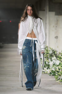 Model on a runway wearing a white oversized blouse with lace tie and distressed blue jeans with a white drawstring belt.