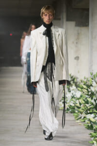 Runway model in a cream pinstripe suit with oversized blazer, black lace necktie, and layered sheer details walking the catwalk in an industrial setting with floral decor nearby.