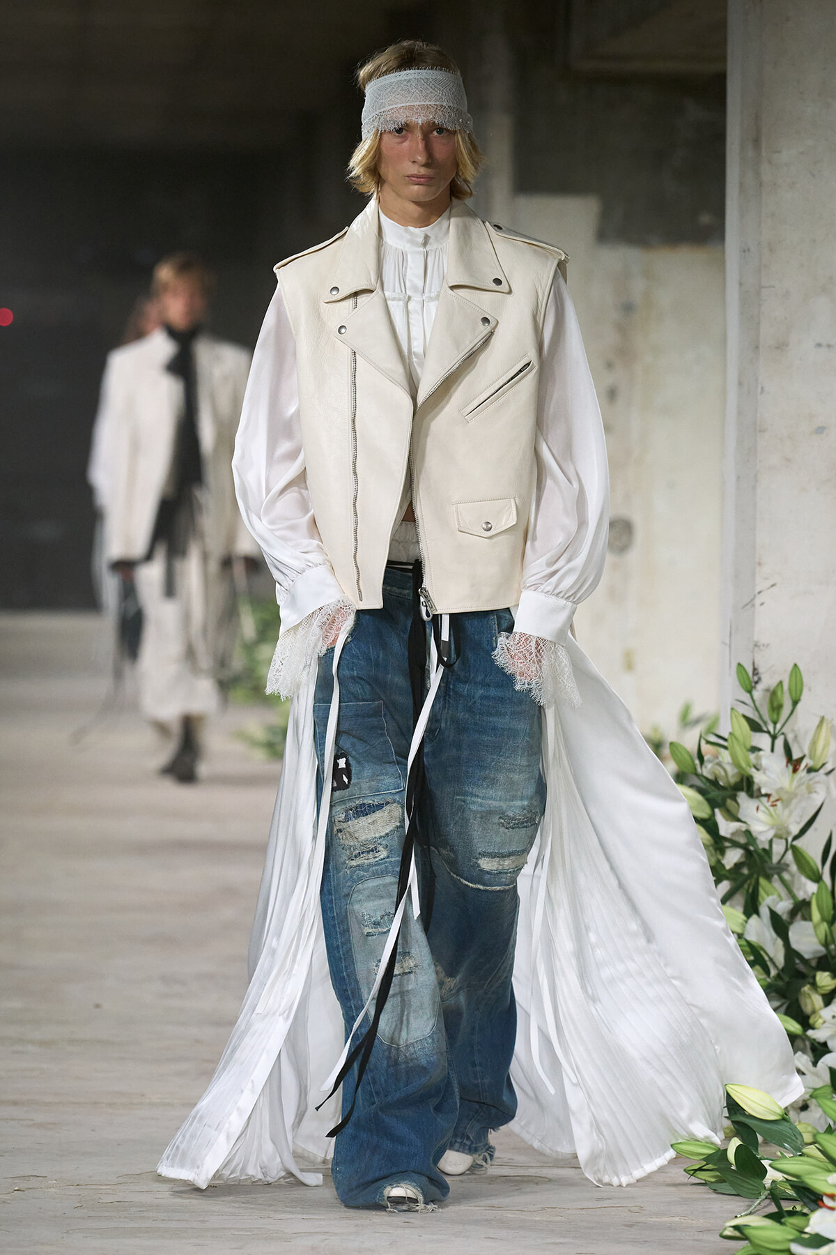 Model on a runway wearing a white vest over a puff-sleeve blouse, lace cuffs, distressed jeans, and a long white cape with a flowing train.
