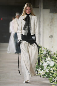 Model walking a runway in a white pinstriped suit with a black lace necktie and layered black wraps at the waist.