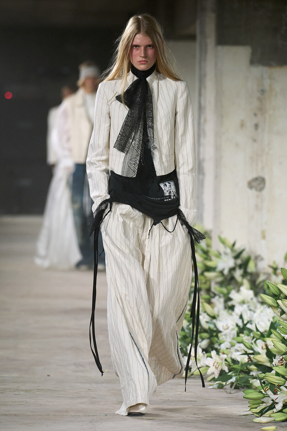Model walking a runway in a white pinstriped suit with a black lace necktie and layered black wraps at the waist.
