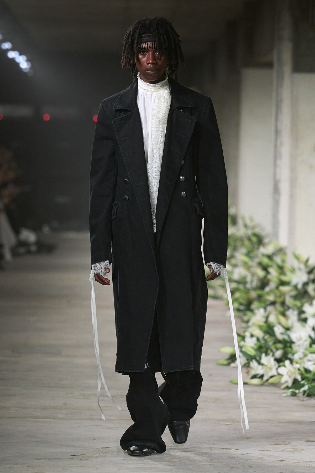 Model on a runway wearing a long black coat over a white lace high-neck blouse, with wide black pants and white sleeve ribbons, in an industrial venue with floral decor along the catwalk.