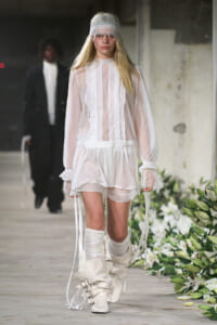 Runway model in a sheer white ruffled dress with lace headband and tall strapped boots walking a fashion show.