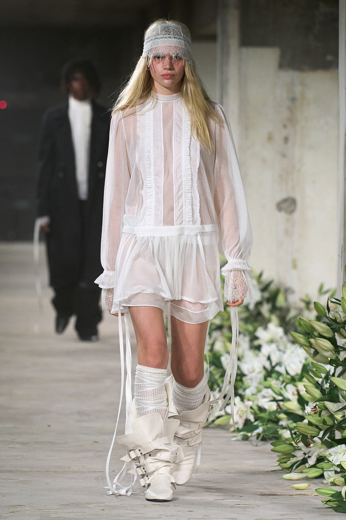 Runway model in a sheer white ruffled dress with lace headband and tall strapped boots walking a fashion show.
