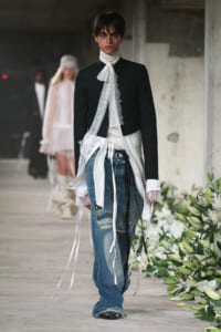 Runway model in a black cropped blazer with a white lace neck scarf and long white ribbons over distressed blue jeans, walking the runway.