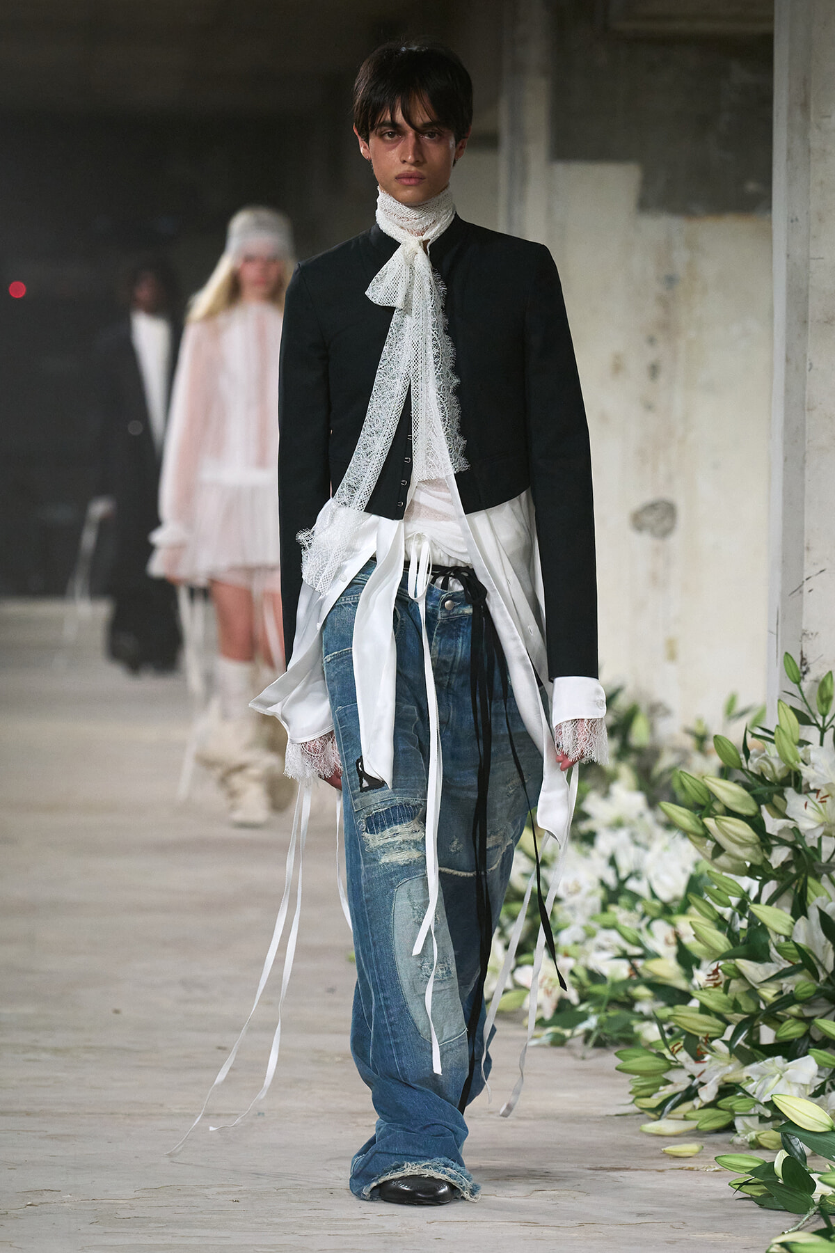 Runway model in a black cropped blazer with a white lace neck scarf and long white ribbons over distressed blue jeans, walking the runway.