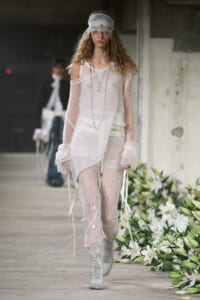 Model walking runway in a sheer white knit top over lace floral pants, layered necklaces, and a fringed headband with silver boots; floral runway decor behind.
