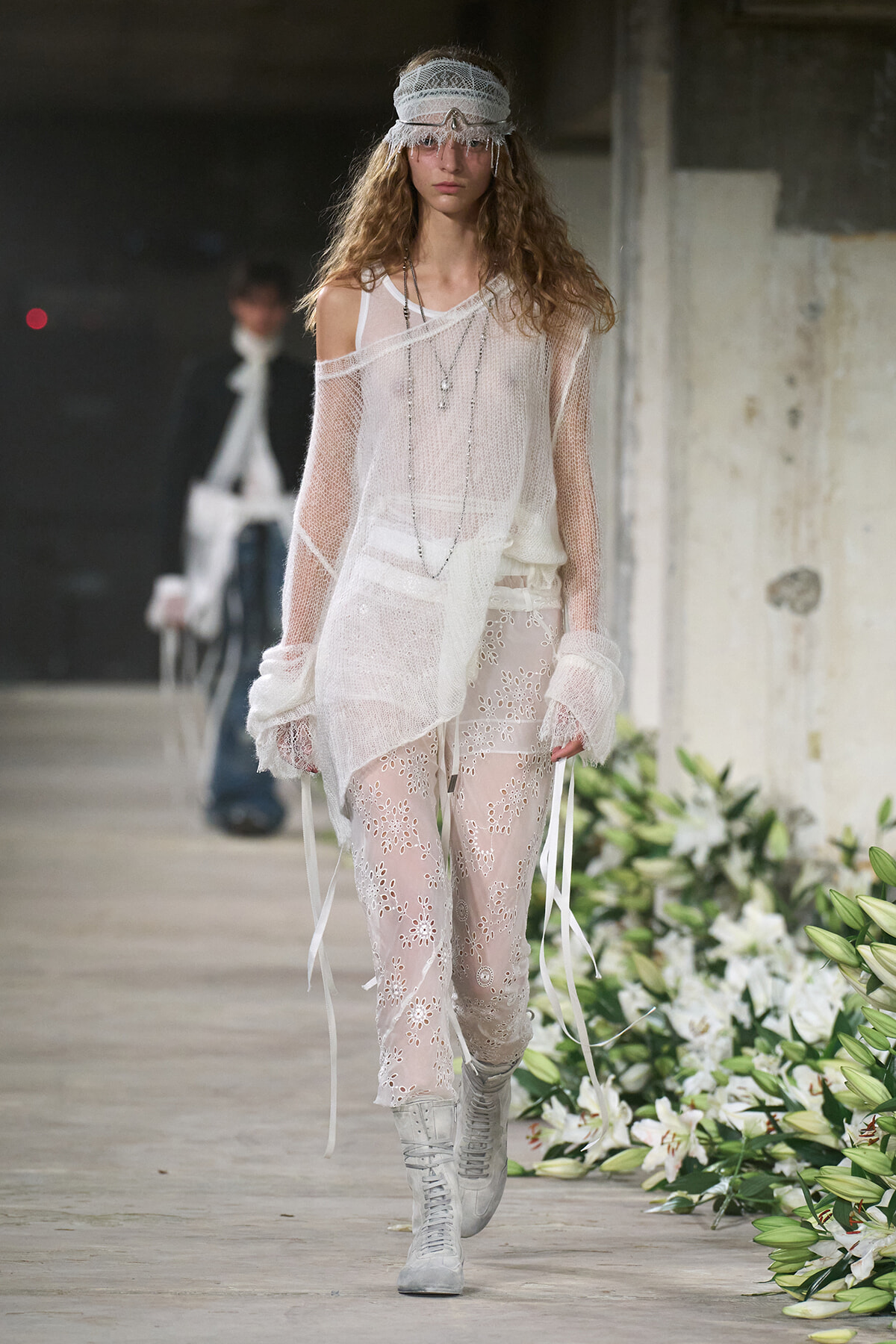 Model walking runway in a sheer white knit top over lace floral pants, layered necklaces, and a fringed headband with silver boots; floral runway decor behind.
