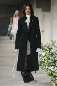 Model on runway wearing a long black double-breasted coat with lace-trim cuffs and a white lace scarf.