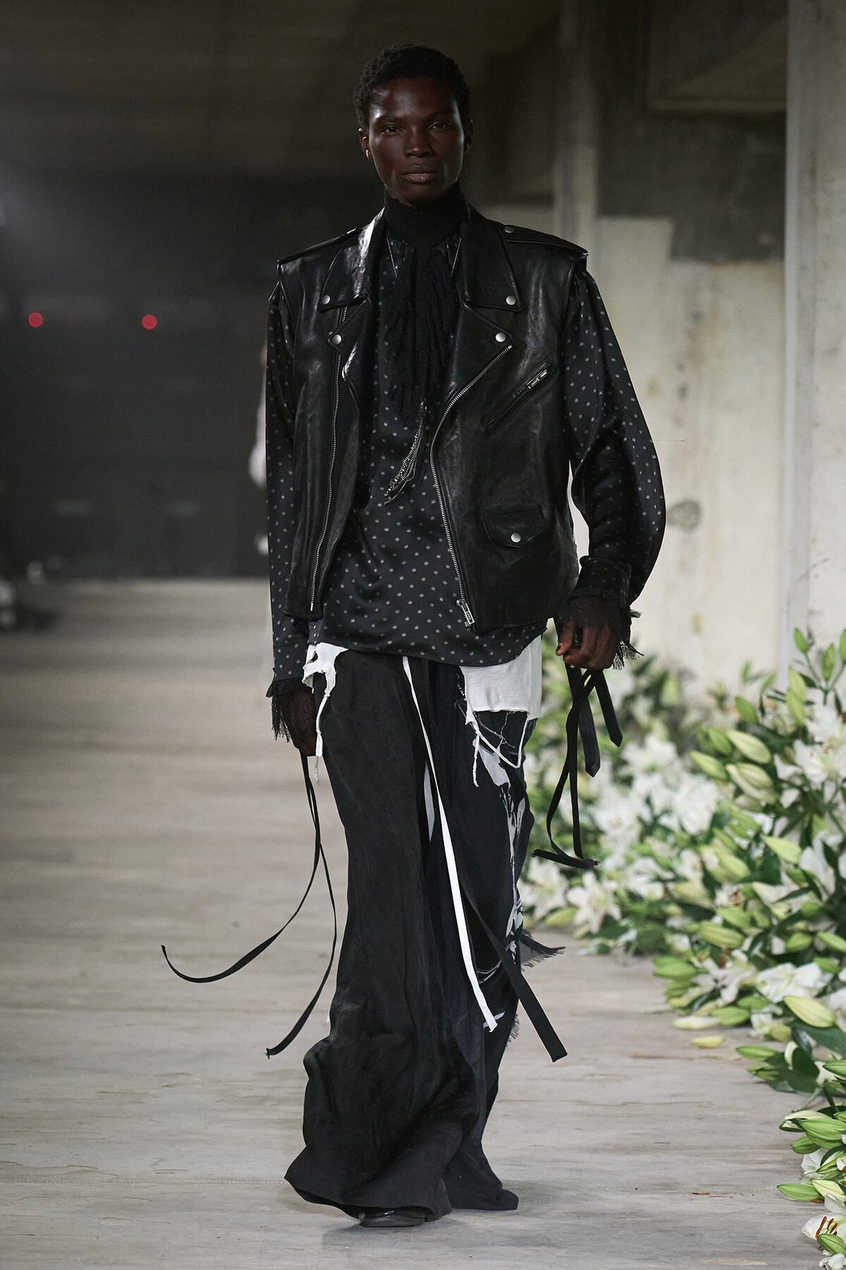 Model walking a runway in a black leather vest over a polka-dot shirt and distressed black-and-white trousers with ribbon details.
