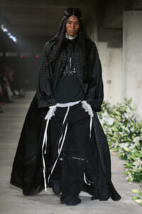 Runway model in an all-black, oversized cape with dotted pattern, layered with lace and ribbons, walking the catwalk.