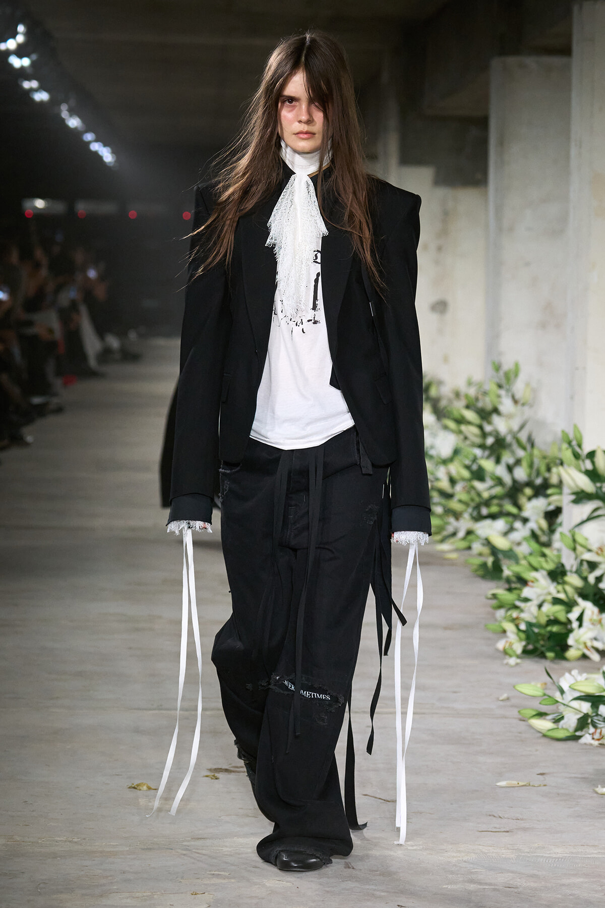 Female model walks a runway wearing a black tailored blazer and pants, with a white lace scarf, in an industrial venue lined with white flowers.