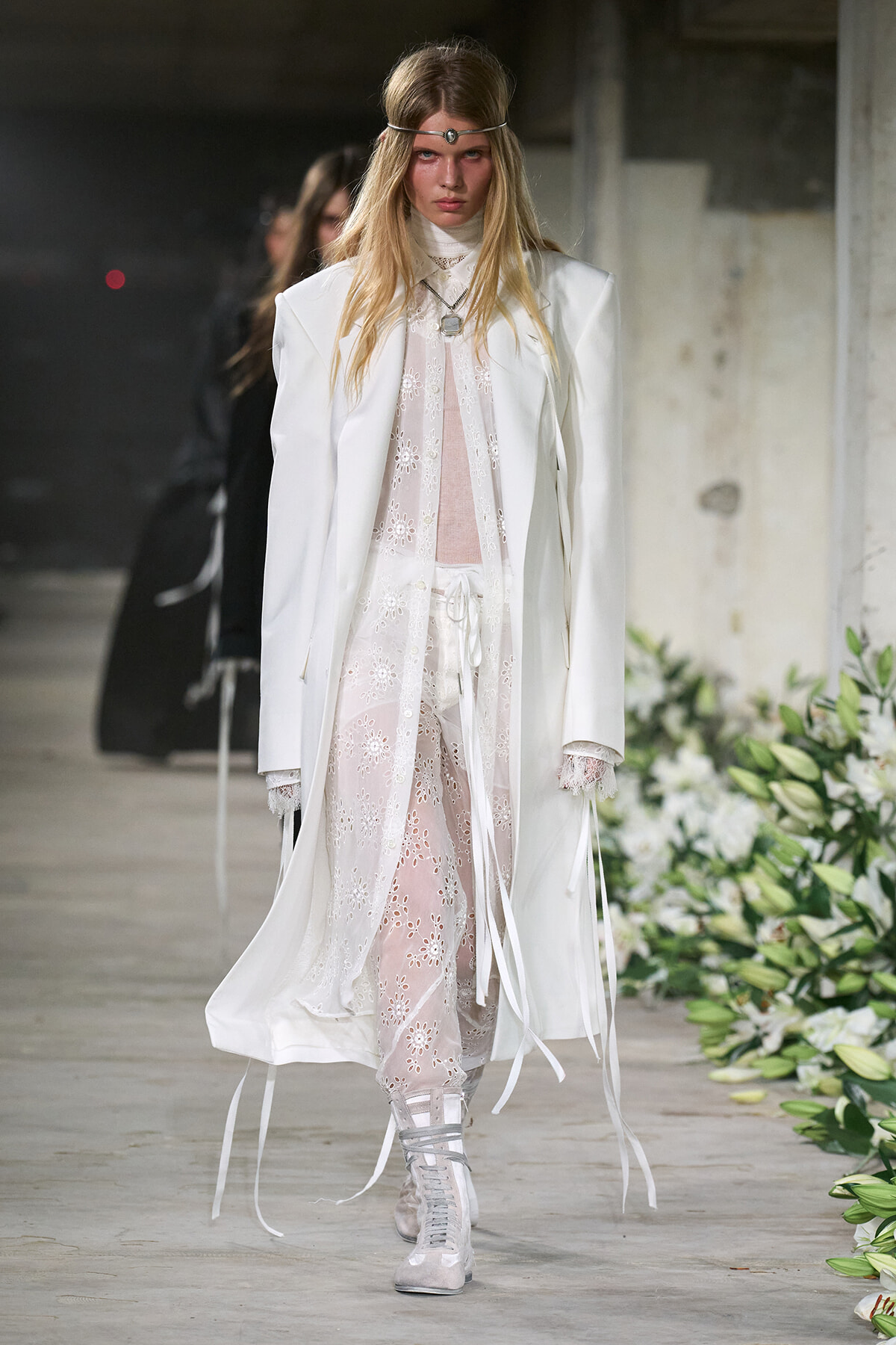 Model walking a runway in a long white coat over sheer floral lace outfits, with silver boots and a slim headband accessory.