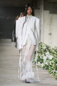Model on a runway in a white oversized blazer over a sheer lace dress, with a silver headband and lavender shoes in an industrial setting with floral arrangements nearby.