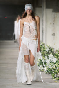 Model walking a runway in a white lace patchwork dress, lace blindfold headband, and white knee-high boots.