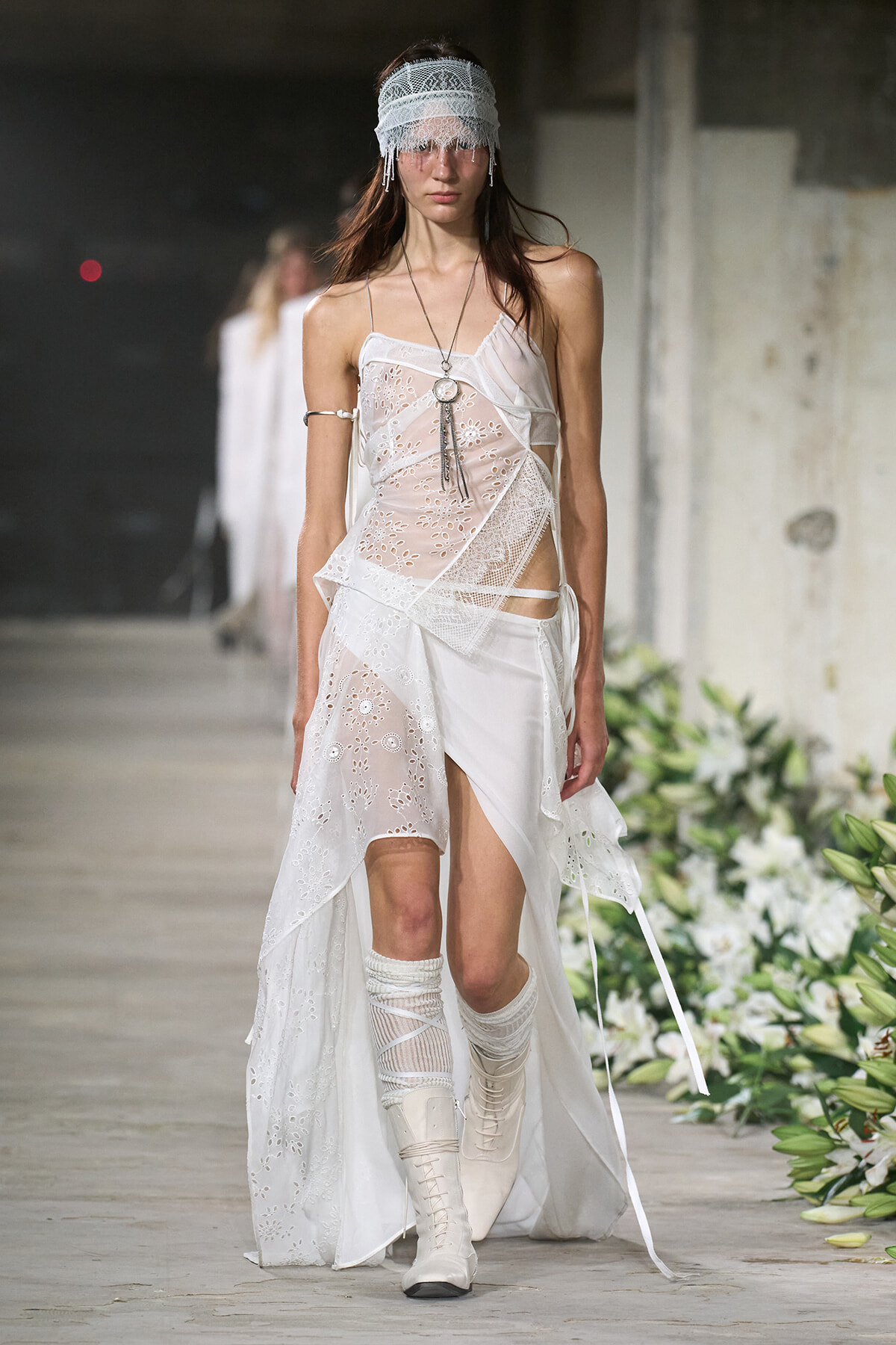 Model walking a runway in a white lace patchwork dress, lace blindfold headband, and white knee-high boots.