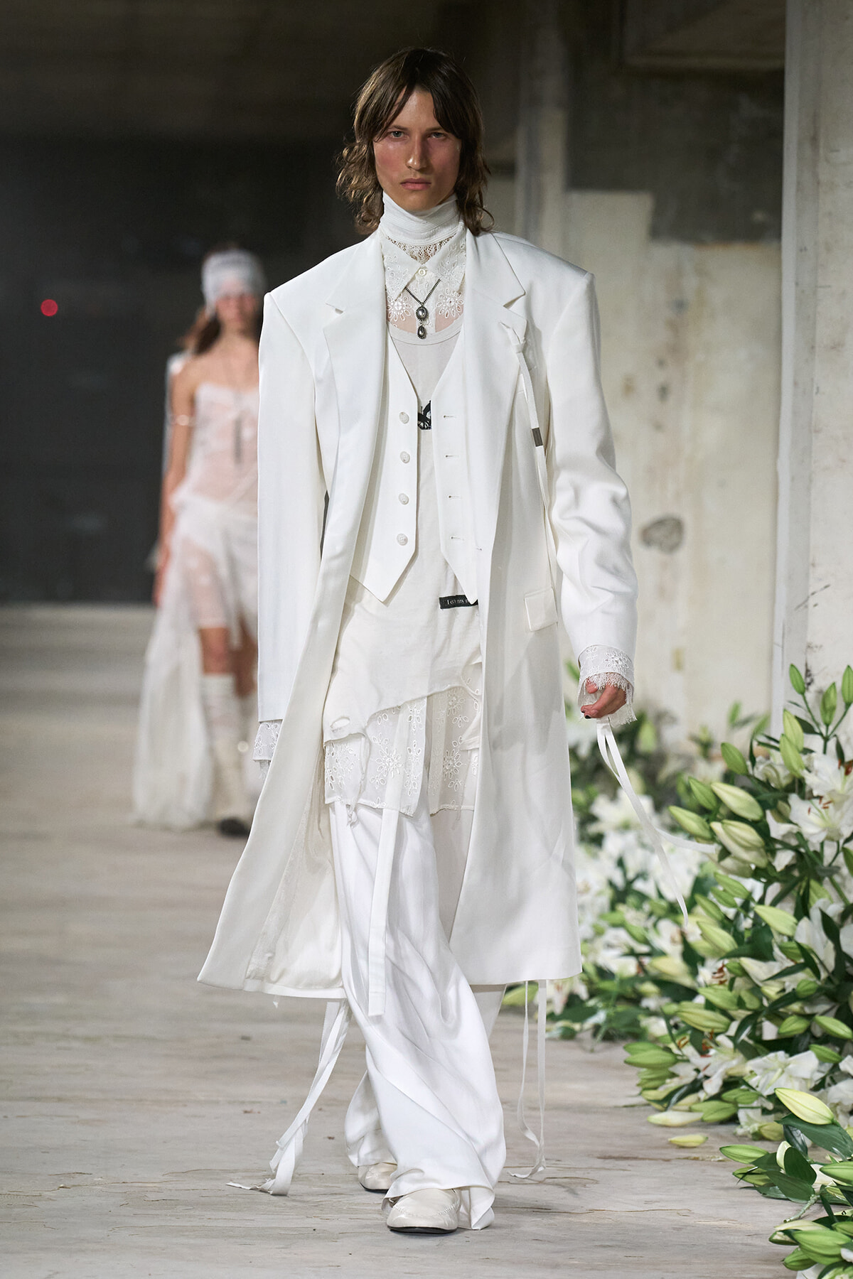 Models white haute couture outfit: long tailored white coat over layered lace top and wide-leg pants, walking a fashion runway.