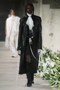 Model walking the runway in a long black leather coat with lace cuffs and a white lace neck scarf, black boots, and layered accessories.