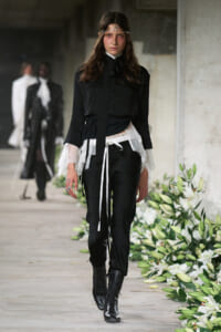 Model walking a runway in a black blouse with lace cuffs, layered white sheer sash, black pants, and tall black boots; a delicate headband sits on her hair.
