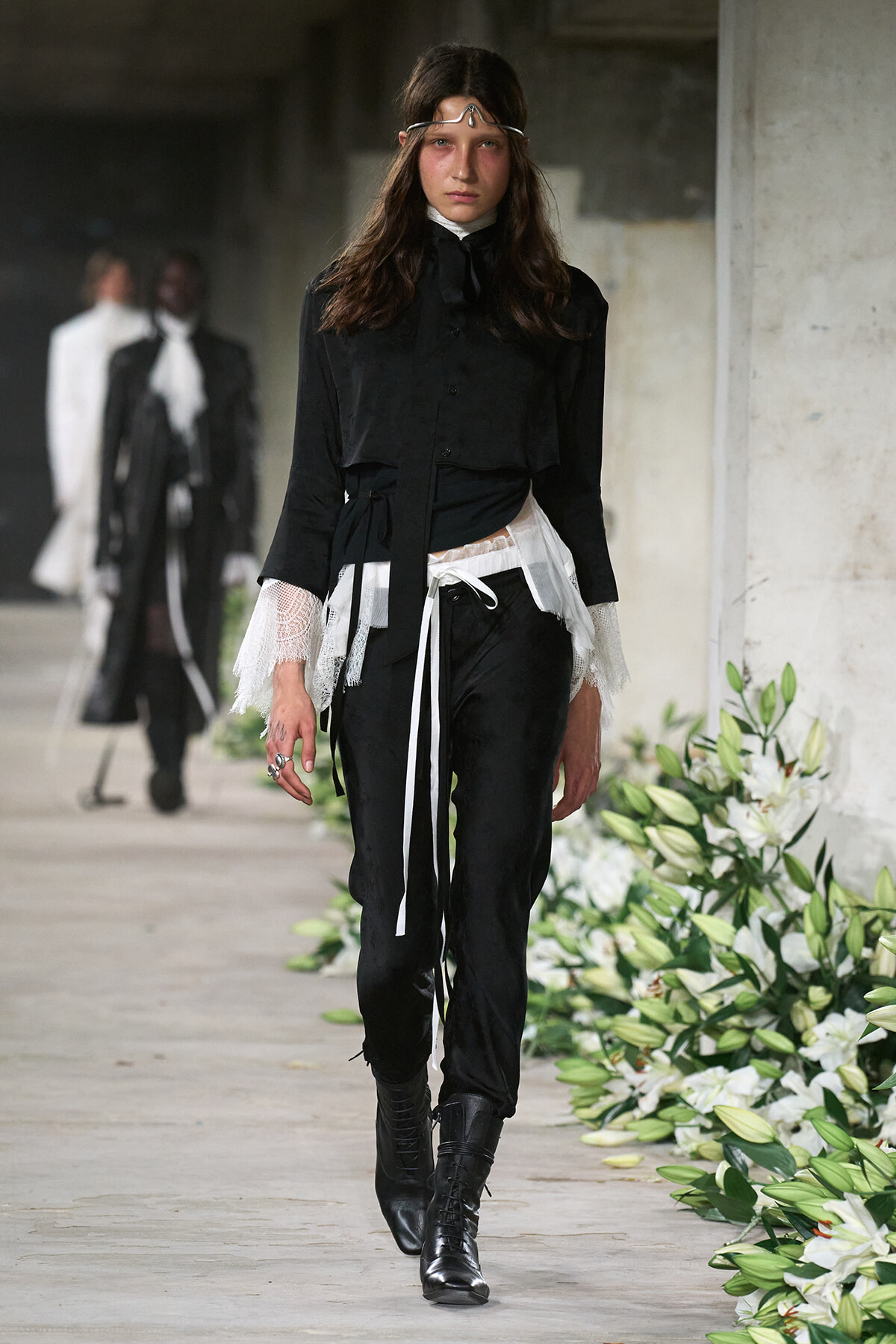 Model walking a runway in a black blouse with lace cuffs, layered white sheer sash, black pants, and tall black boots; a delicate headband sits on her hair.