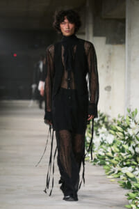 Male model walks a fashion runway in an all-black sheer lace outfit with a vest and long ribbon details.