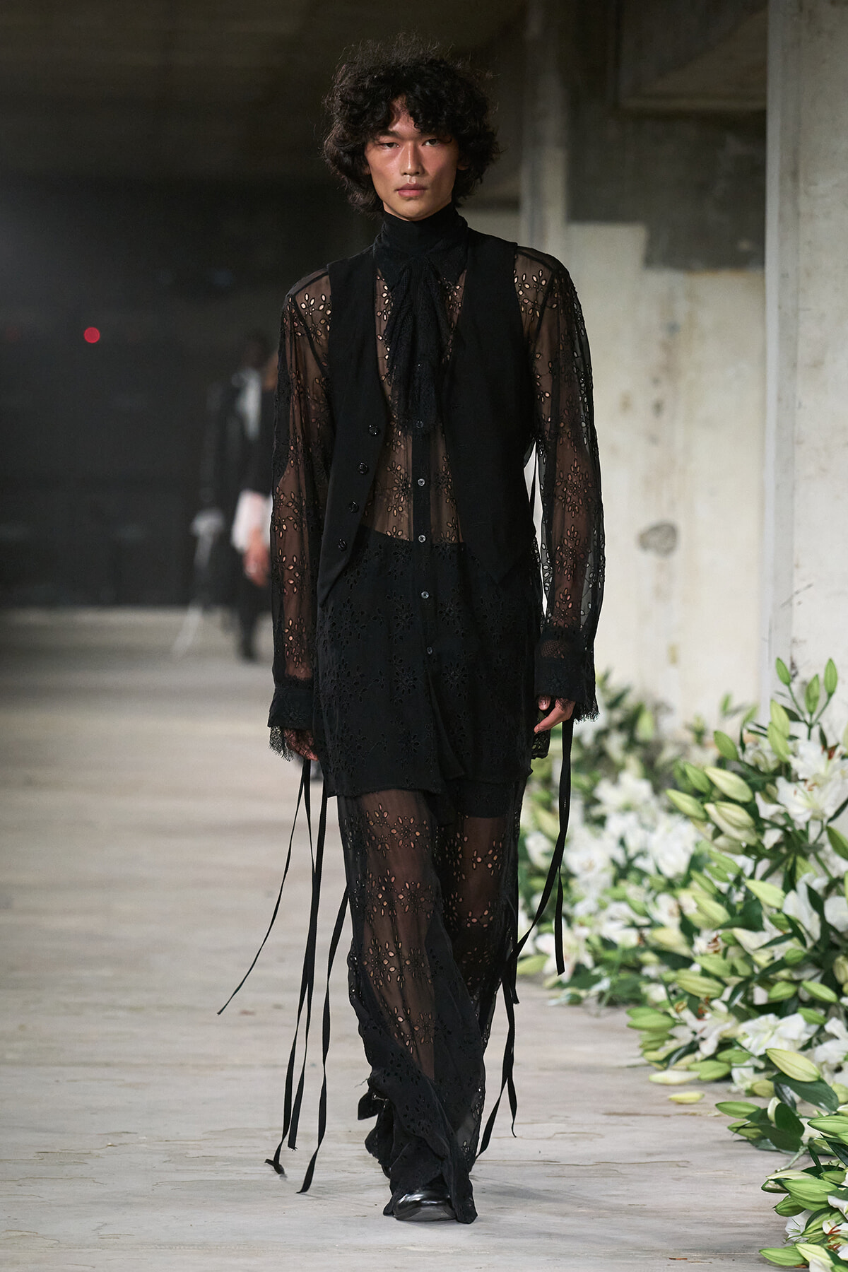 Male model walks a fashion runway in an all-black sheer lace outfit with a vest and long ribbon details.